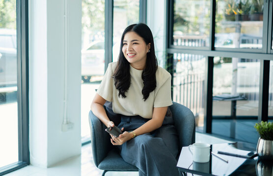 Happy Asian businesswoman holding smartphone sitting in armchair during coffee break near window inside modern corporate office background