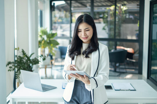 Confident Asian businesswoman using digital tablet with stylus pen standing near desk in modern corporate office background