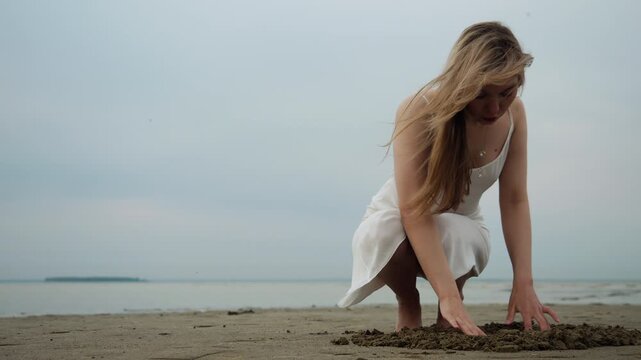 Young woman in a white dress crouches barefoot on a quiet beach and touches the sand with her hands near the water, enjoying a peaceful moment outdoors.