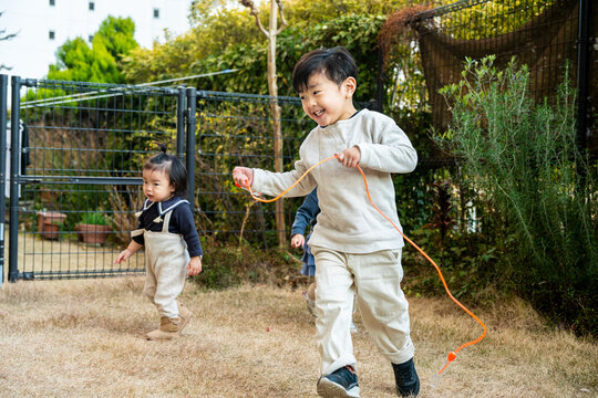 A boy skipping rope and running outdoors with a toddler walking nearby.