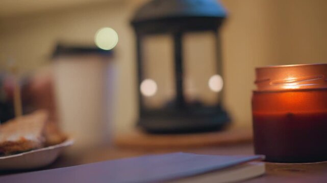 Close up of lit candle in glass jar on wooden table with soft warm light and bokeh background. Notebook lies nearby suggesting quiet workspace or study environment. Candle burns steadily