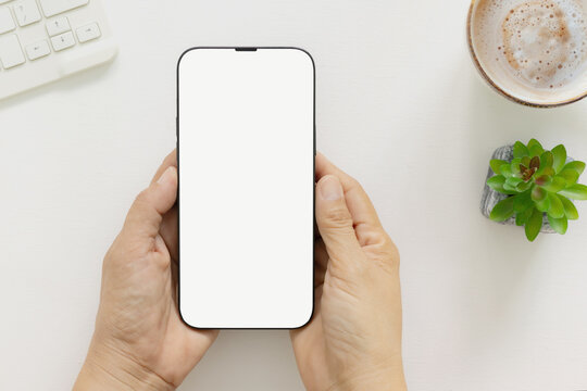 Top view of hand holding modern smartphone with blank white screen template and keyboard and small green plant, coffee on white desk. Concept for marketing, website, branding, social.