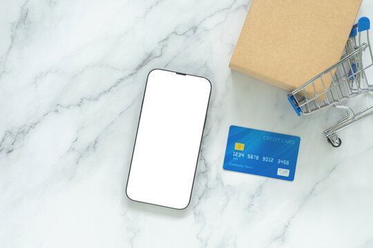 Blank smartphone mockup with blue credit card, shopping cart and kraft package on white marble, Concept ecommerce, online shopping, mobile payment, retail, digital commerce and product promotion.