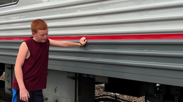 The teenager is standing on the platform, leaning on the train with his hand, and waiting to board the car.