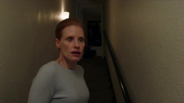 Scared woman with a shocked expression turns around and runs up a dark, narrow staircase in a dimly lit hallway, creating a suspenseful horror scene.