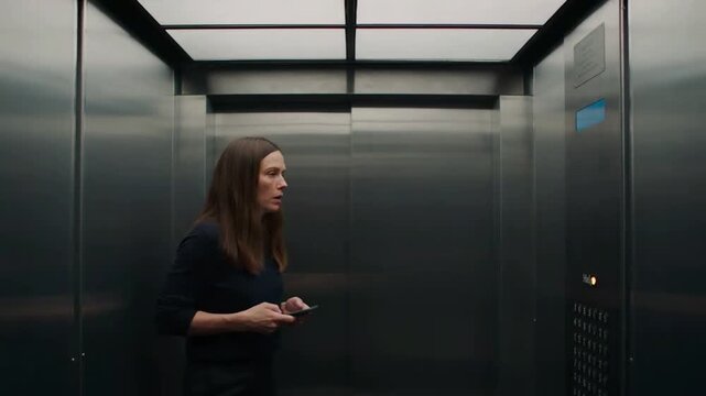 A frightened woman alone in a dark modern metallic elevator looking up with a worried expression while holding a smartphone in a suspenseful scene.