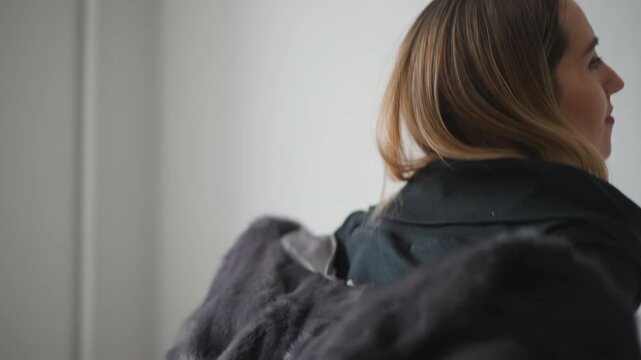Close up of woman putting on and adjusting black fur coat in front of plain white wall. subject is seen from side as woman lifts drapes and secures garment over her shoulders revealing its