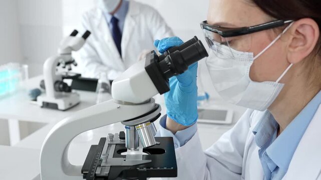 Masked professional female scientist examining sample through microscope, conducting research alongside colleague in sterile laboratory setting. Medicine, healthcare and science concept