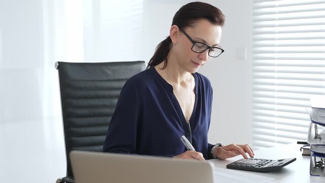 Professional female wearing black glasses, is working at desk, calculating finances, writing notes, managing business documents in contemporary workspace. Audit and taxes in business