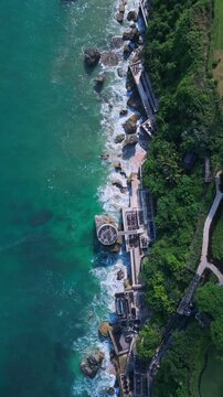 Aerial view cliff restaurant jimbaran bali, rocky coastline with turquoise water, terraced dining platforms and crashing waves, lush tropical foliage, chefs arranging tables
