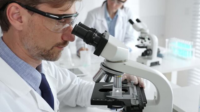 Researcher examining sample through microscope, wearing safety eyewear in sterile laboratory setting, surrounded by scientific glassware. Medicine, healthcare and science concept