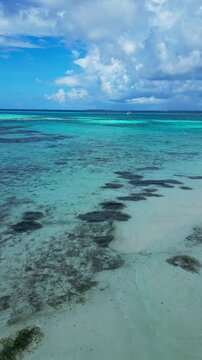 Sunlit turquoise water shows clear reef textures and shallow sandy seabed near shore, while a distant boat rests on the horizon under bright clouds in tropical scene.Vertical video