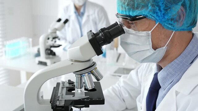 Senior male scientist wearing a protective mask, cap, and gloves, examining samples under a microscope in a modern laboratory. Medicine, healthcare and science concept