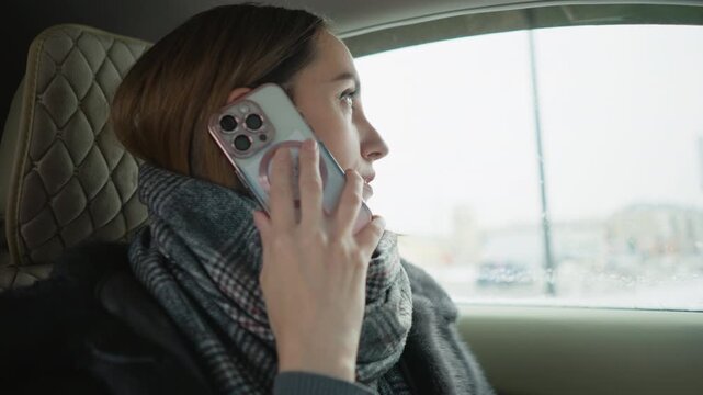 Close up of woman talking on smartphone while seated in car seat with quilted headrest. Woman wears gray scarf and dark coat holding phone to ear during call. Bright light from window