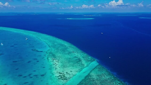 Aerial view of turquoise lagoon blending into deep blue ocean over a vivid coral reef with small boats drifting across clear water under a sunny sky with clouds.