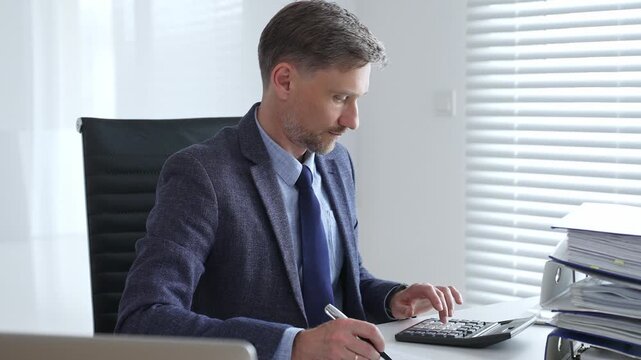 Professional businessman calculating financial data, wearing business attire while reviewing paperwork at sleek workplace desk. Audit and taxes in business