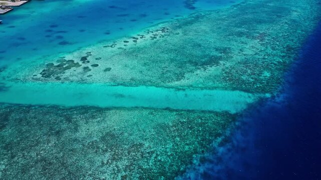 Aerial view of turquoise lagoon blending into deep blue ocean over a vivid coral reef with small boats drifting across clear water under a sunny sky with clouds.