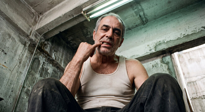 Contemplative older man sitting in a rundown industrial space, resting his chin on his hand under harsh fluorescent light. Raw, emotional character study.