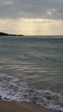 A serene beach scene with gentle waves lapping at the shore. The sky is overcast, creating a calm atmosphere. The ocean reflects soft light.