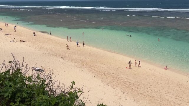 A sandy beach with clear turquoise water. People of various ages and ethnicities walk along the shore. Gentle waves lap at the sand under a blue sky.