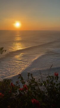A serene sunset over the ocean, with gentle waves and vibrant flowers in the foreground. The sky is painted in warm hues of orange and yellow.