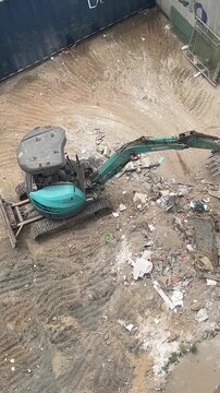 Aerial view of a construction site with a green excavator on a dirt surface. Debris and rubble scattered around the area.