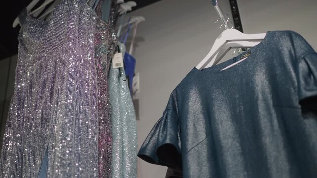 Close up of various sparkly dresses and garments hanging on hangers in retail setting. clothing features shiny sequins glitter details and different textures under soft lighting. Background