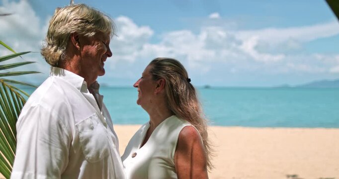 Senior couple standing closely under palm tree leaves on a sunny beach in Samui, Thailand, gazing affectionately at each other, celebrating love and romance during their tropical resort vacation