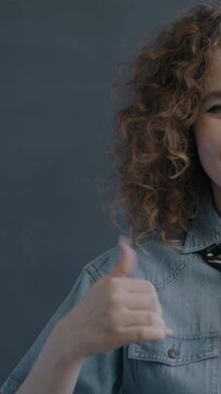 Portrait of happy young woman showing thumbs-up hand gesture and winking on black studio background. People and positive emotion concept.