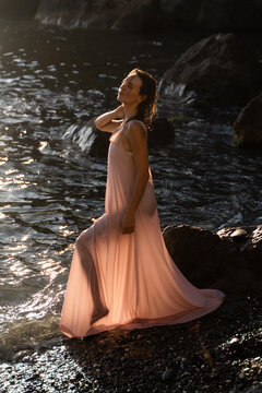 Serene young woman in light pink dress posing peacefully in shallow coastal waters during sunset
