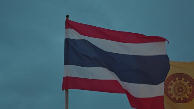 National flag of thailand and a yellow buddhist flag fluttering against a blue evening sky