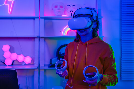 Young woman wearing VR headset and holding controllers playing virtual reality game at home