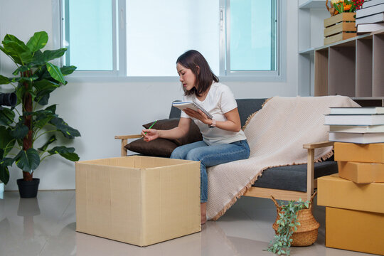 Asian woman small business owner taking inventory notes with cardboard boxes at home office