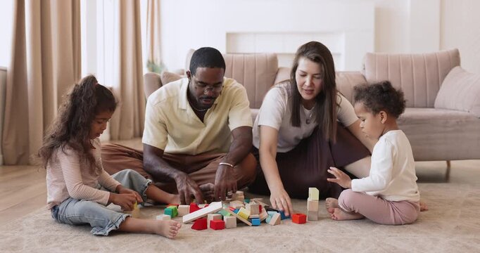 Happy interracial parents constructing toy city from wooden blocks with two little girls on floor in living room teaching children through creative building game supporting early childhood development