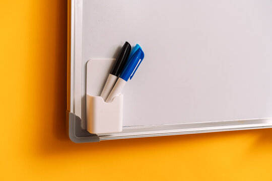 Black and blue markers in a holder at a whiteboard corner beside a yellow wall.