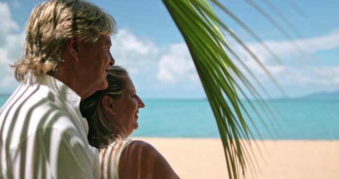 Mature couple relaxing on a tropical beach vacation in Thailand, looking out at the turquoise ocean and pointing into the distance from behind a palm leaf. Romantic getaway on Samui Island. Close up