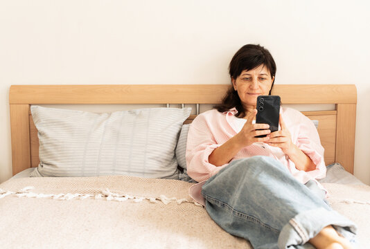 Mature woman relaxes in bed at home, wearing pink shirt and jeans, looks at her smartphone with gentle smile, scrolling through social media and enjoying peaceful morning break, digital connections