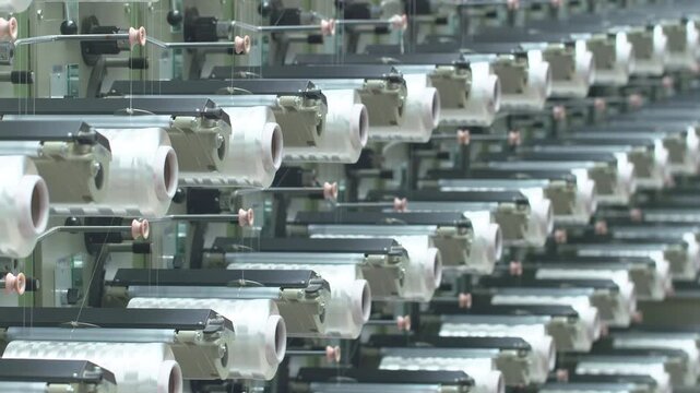 Industrial factory production line of weaving plant of plastic or polypropylene bags yarn industrial. Rows of white reels with ropes making plastic sacks in a modern nylon manufacture enterprise