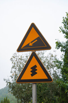 Orange triangular road signs warning of a steep slope and sharp turn ahead on a mountain road, concept of life challenges, risk, caution and difficult journey ahead