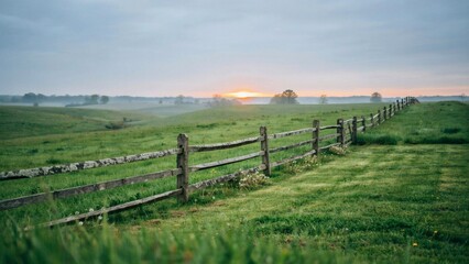 Naklejka premium A weathered wooden split rail fence stretches across a green misty meadow at sunrise hour with rolling meadow landscape divides grassy rolling