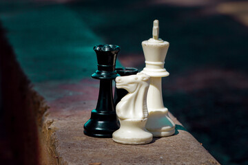 Close-up shot of black and white chess pieces including king, queen, and knight standing together on an outdoor stone ledge. © Xihy