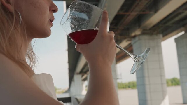 Woman holds and drinks red wine under bridge with industrial structure in background. scene features soft natural light and outdoor urban setting details