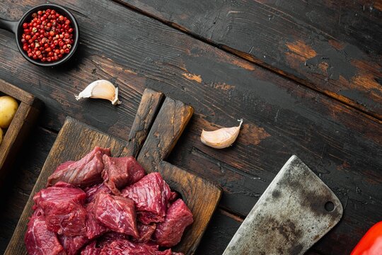 Chopped raw stewing beef cubes arranged on a dark weathered wooden cutting board beside a metal cleaver, scattered garlic cloves and a bowl of red peppercorns on a rustic dark wood table
