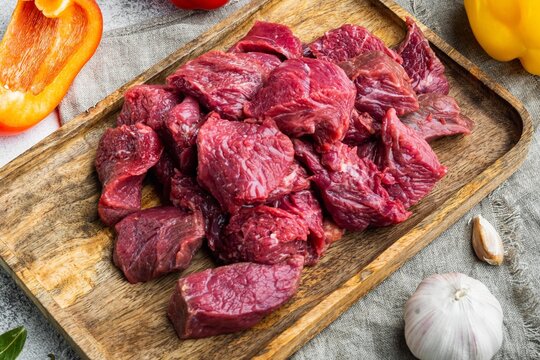 Chopped raw stewing beef chunks arranged on a rustic wooden cutting board beside whole garlic bulb and halved bell peppers under bright natural light