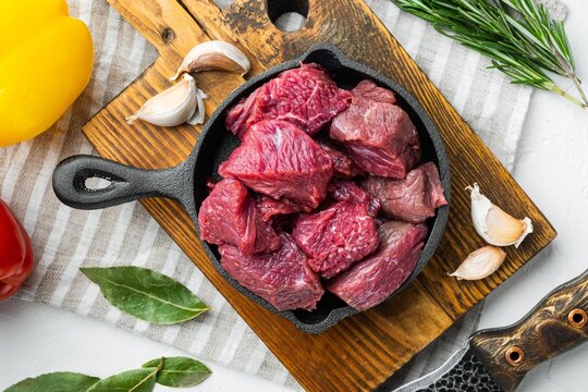 Raw diced beef cubes in a black cast iron skillet resting on a rustic wooden cutting board beside whole garlic cloves fresh rosemary sprigs and green bay leaves with yellow