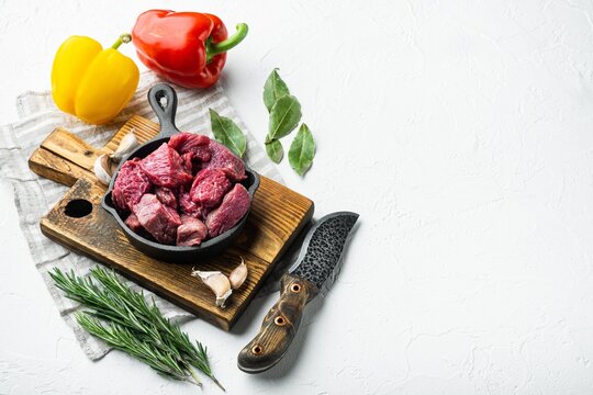 Cast iron skillet filled with diced raw stewing beef cubes arranged on a rustic wooden cutting board beside red and yellow bell peppers, fresh bay leaves, rosemary sprigs, whole garlic cloves
