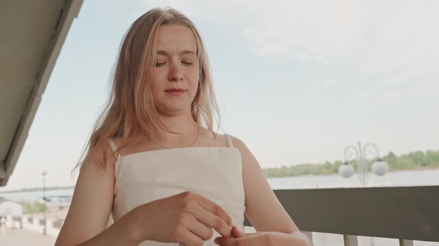 Woman in white dress holds and examines measuring tape on outdoor balcony with water trees sky background. Woman manipulates fabric of her sleeve while holding or adjusting measurement tool