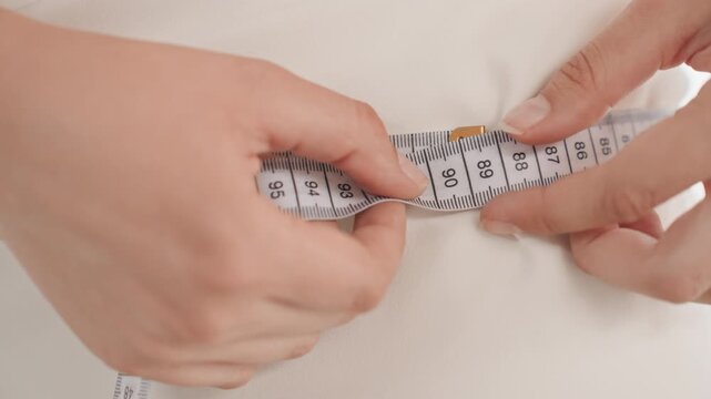 Woman measures her body with white measuring tape focusing on torso area. close up view shows hands adjusting and reading numbers on flexible fabric ruler against smooth skin. Soft lighting
