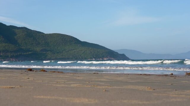 Captivating Shoreline: A Serene Beach Scene Highlighting Gentle Waves, Sandy Coast, and Lush Green Hills Under a Clear Blue Sky
