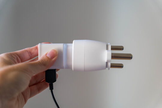 Hand holds a travel adapter with three round pins of the South African plug standard and a connected charging cable. Electrical compatibility for travelers packing gear for a trip abroad.